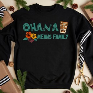 May include: Black sweatshirt with the text "OHANA MEANS FAMILY" in teal and a tiki design. The design includes orange, yellow, and purple flowers. The sweatshirt is surrounded by wrapped gifts and pine branches.