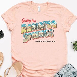 K&ouml;nnte beinhalten: Ein rosa T-Shirt mit einem Vintage-Postkarten-Design mit dem Text "Greeting from Radiator Springs, Gateway to the Ornament Valley".
