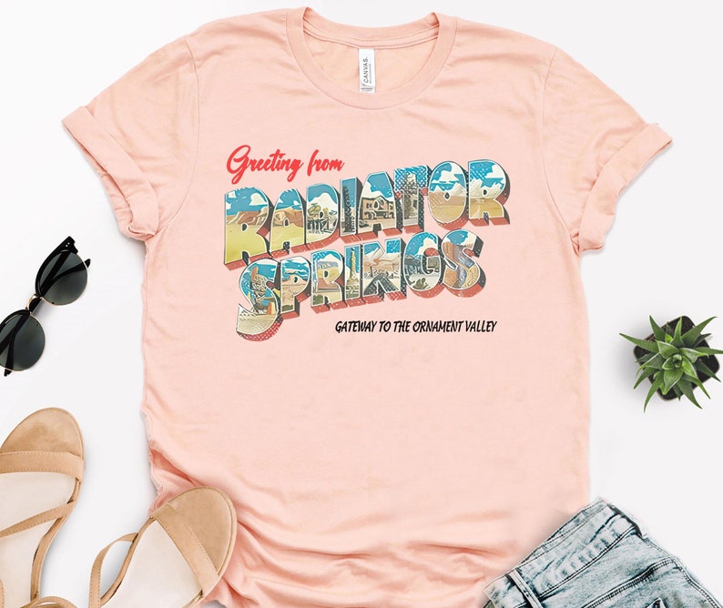 K&ouml;nnte beinhalten: Ein rosa T-Shirt mit einem Vintage-Postkarten-Design mit dem Text "Greeting from Radiator Springs, Gateway to the Ornament Valley".