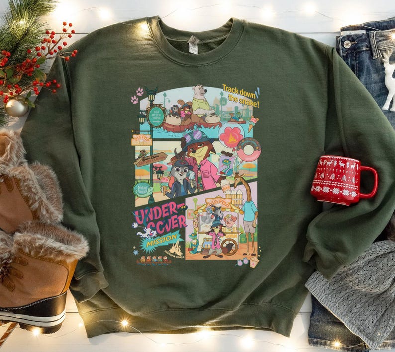 May include: Olive green sweatshirt with a colorful graphic featuring cartoon animals and the text "Undercover Mission." The graphic includes the phrase "Track down the snake!" A red mug with a white holiday design is visible.