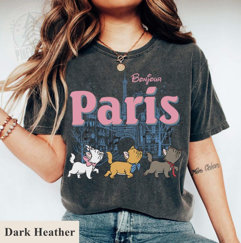 Puede incluir: Camiseta gris jaspeado oscuro con la frase "Bonjour Paris" en rosa, una ilustraci&oacute;n de la Torre Eiffel y tres gatos de dibujos animados. Los gatos son blancos, marrones y grises, cada uno con un lazo.