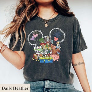 Pu&ograve; includere: T-shirt grigio scuro melange con una grafica dei personaggi di Toy Story della Disney all'interno di un contorno della testa di Topolino. Il design include Buzz Lightyear, Woody e altri personaggi. Il testo "Dark Heather" &egrave; in basso.