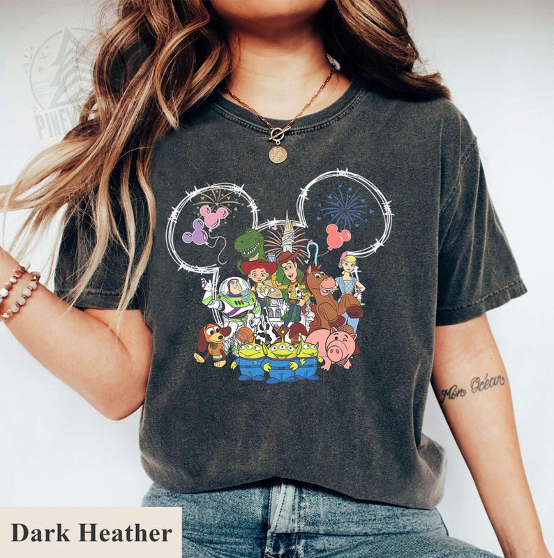 Pu&ograve; includere: T-shirt grigio scuro melange con una grafica dei personaggi di Toy Story della Disney all'interno di un contorno della testa di Topolino. Il design include Buzz Lightyear, Woody e altri personaggi. Il testo "Dark Heather" &egrave; in basso.