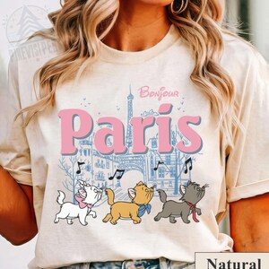 Puede incluir: Camiseta beige con la palabra "Paris" en rosa, una ilustraci&oacute;n azul de la Torre Eiffel y edificios, y tres gatos de dibujos animados cantando con notas musicales. La palabra "Bonjour" est&aacute; encima de "Paris".