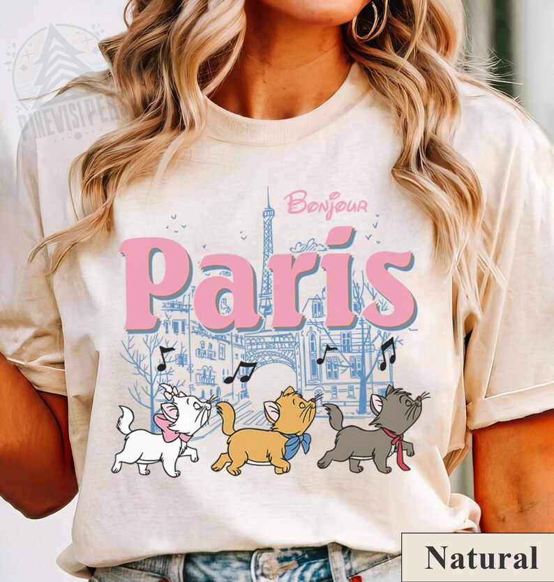 Puede incluir: Camiseta beige con la palabra "Paris" en rosa, una ilustraci&oacute;n azul de la Torre Eiffel y edificios, y tres gatos de dibujos animados cantando con notas musicales. La palabra "Bonjour" est&aacute; encima de "Paris".