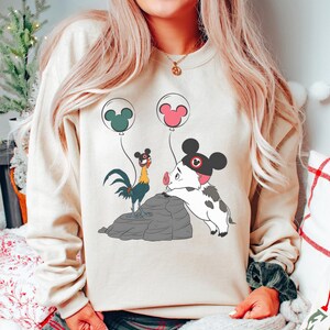 May include: Beige crew neck sweatshirt with a cartoon illustration of a rooster, a pig, and two balloons with Mickey Mouse ears. The pig is wearing a black hat with a red Mickey Mouse emblem. The rooster is standing on a grey rock.