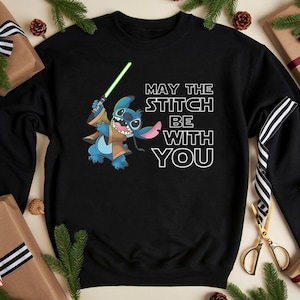 May include: Black sweatshirt featuring a blue cartoon character holding a green lightsaber, with the text "MAY THE STITCH BE WITH YOU". The design includes brown and tan accents. The sweatshirt is surrounded by wrapped gifts and pine branches.