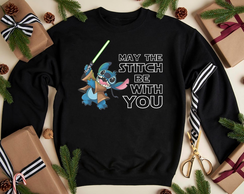May include: Black sweatshirt featuring a blue cartoon character holding a green lightsaber, with the text "MAY THE STITCH BE WITH YOU". The design includes brown and tan accents. The sweatshirt is surrounded by wrapped gifts and pine branches.
