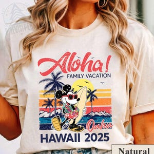 May include: A natural-colored t-shirt featuring a colorful graphic of Mickey Mouse in sunglasses and a Hawaiian shirt, holding a surfboard. The design includes the words "Aloha! Family Vacation" and "Hawaii 2025" with a sunset and palm trees.