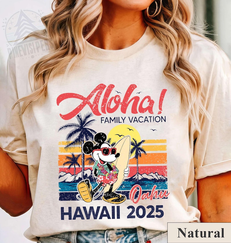 May include: A natural-colored t-shirt featuring a colorful graphic of Mickey Mouse in sunglasses and a Hawaiian shirt, holding a surfboard. The design includes the words "Aloha! Family Vacation" and "Hawaii 2025" with a sunset and palm trees.
