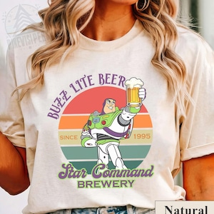 May include: Beige t-shirt featuring a graphic of Buzz Lightyear holding a beer, with the text "Buzz Lite Beer" and "Star Command Brewery." The design includes a retro sunset background and the year "1995."