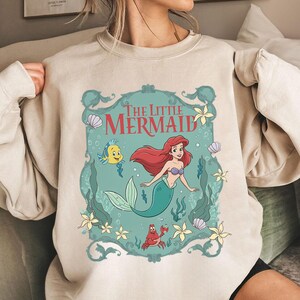 May include: Beige sweatshirt featuring a colorful graphic of Ariel from The Little Mermaid. The design includes Ariel with red hair and a teal tail, along with Flounder and Sebastian. The words "The Little Mermaid" are at the top.