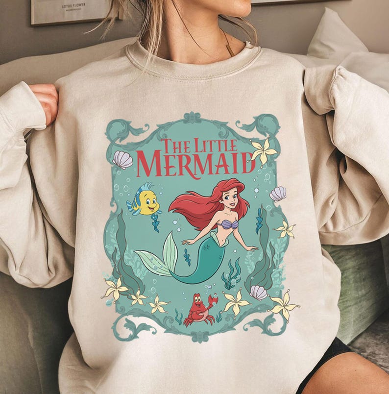 May include: Beige sweatshirt featuring a colorful graphic of Ariel from The Little Mermaid. The design includes Ariel with red hair and a teal tail, along with Flounder and Sebastian. The words "The Little Mermaid" are at the top.