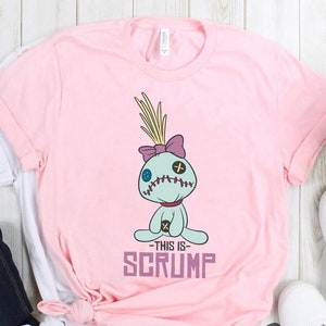 May include: Pink t-shirt with a cartoon image of a blue and green character with a purple bow and the text "This is Scrump" printed on the front.