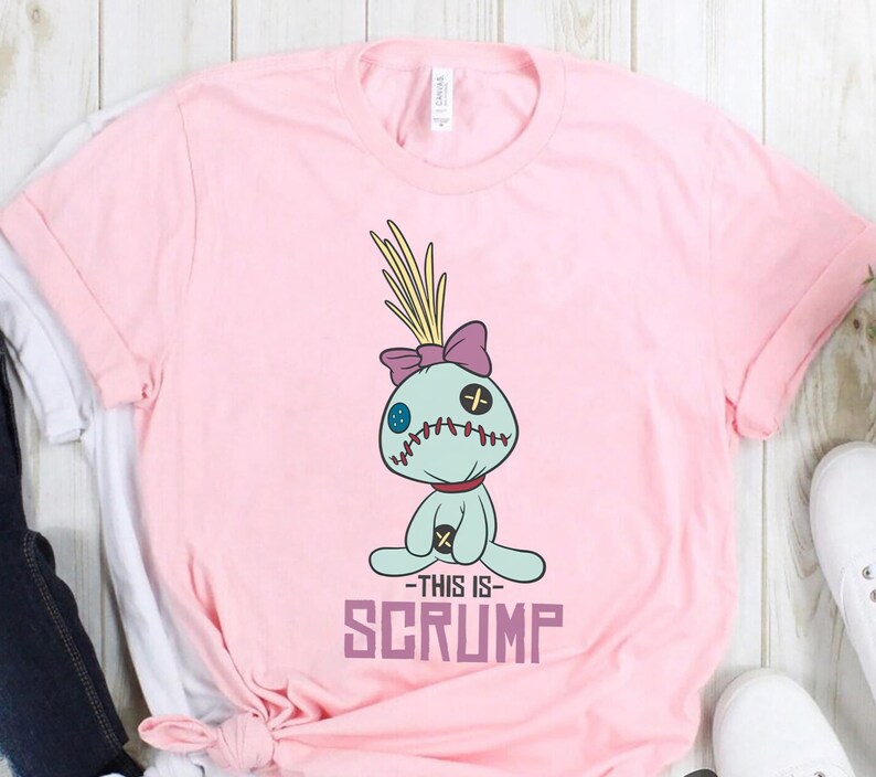 May include: Pink t-shirt with a cartoon image of a blue and green character with a purple bow and the text "This is Scrump" printed on the front.