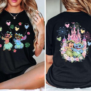May include: Black t-shirt featuring a colorful graphic of two cartoon characters holding Mickey Mouse balloons. The front design shows the characters in sunglasses and grass skirts. The back design shows a castle and fireworks.
