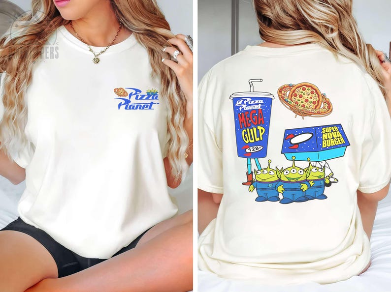 May include: Cream-colored t-shirt featuring the Pizza Planet logo on the front. The back displays a Mega Gulp cup, pizza, a Super Nova Burger box, and three green aliens in blue jumpsuits. A fun, cartoon-style design.