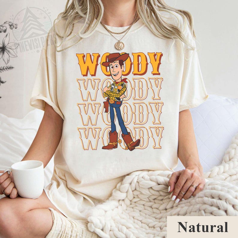 Puede incluir: Camiseta color crema con la palabra "WOODY" en naranja y una imagen de dibujos animados de Woody de Toy Story. El dise&ntilde;o se repite varias veces. La camiseta es de corte holgado.