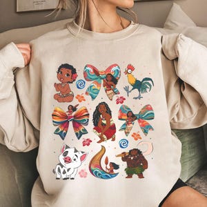 May include: Beige sweatshirt featuring colorful illustrations of characters and elements from a popular animated film. The design includes images of a baby, a rooster, a pig, and a woman, along with decorative bows and floral accents.