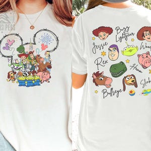 Pu&ograve; includere: T-shirt bianca con una grafica colorata dei personaggi di Toy Story sul davanti, all'interno di un contorno della testa di Topolino. Il retro mostra ritratti di personaggi e nomi, come Woody e Buzz.