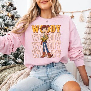 Puede incluir: Una sudadera rosa claro con un gr&aacute;fico del personaje Woody de Toy Story. La palabra "WOODY" est&aacute; impresa en una fuente retro encima y debajo de la ilustraci&oacute;n del personaje. La sudadera tiene mangas largas y cuello redondo.