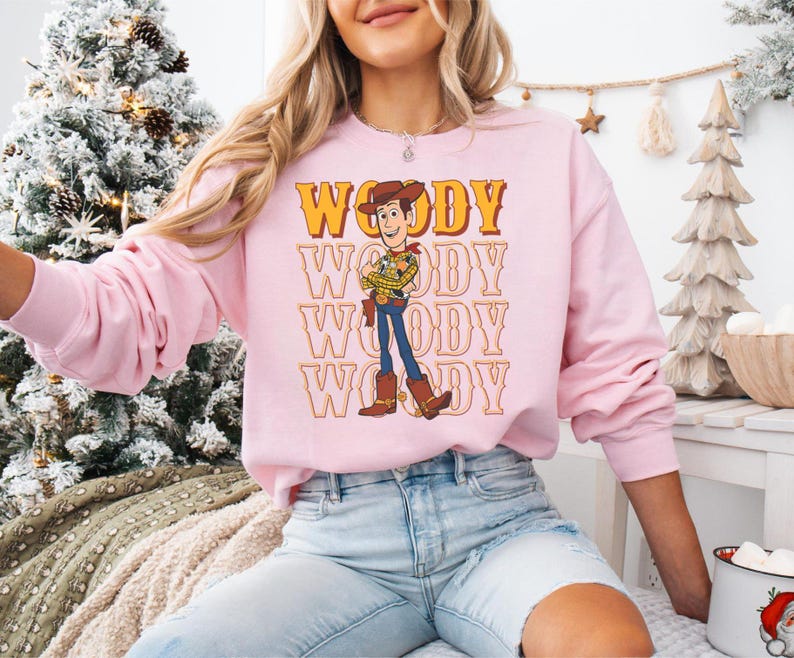 Puede incluir: Una sudadera rosa claro con un gr&aacute;fico del personaje Woody de Toy Story. La palabra "WOODY" est&aacute; impresa en una fuente retro encima y debajo de la ilustraci&oacute;n del personaje. La sudadera tiene mangas largas y cuello redondo.