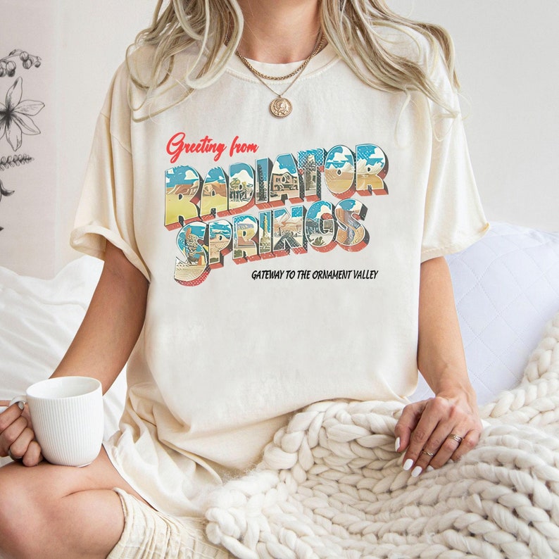 K&ouml;nnte beinhalten: Ein cremefarbenes T-Shirt mit einer Grafik im Vintage-Stil, auf dem "Greeting from Radiator Springs, Gateway to the Ornament Valley" steht.