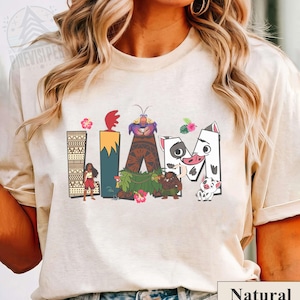 May include: Beige t-shirt with the word "IAM" in a colourful, cartoon-style design. The design features characters and elements from a popular animated film, including a volcano, flowers, and a pig.