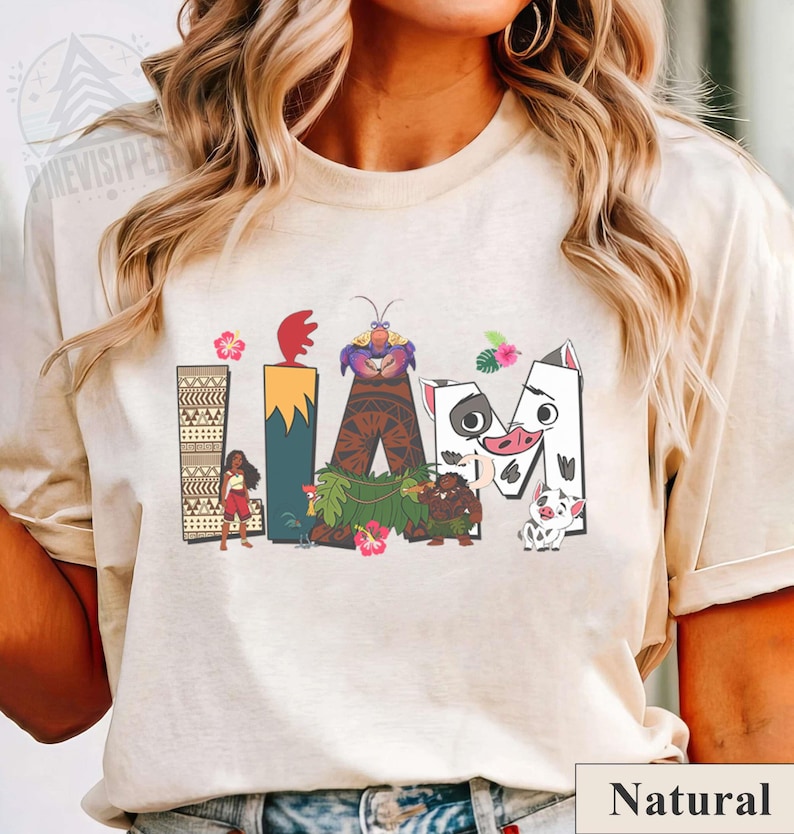 May include: Beige t-shirt with the word "IAM" in a colourful, cartoon-style design. The design features characters and elements from a popular animated film, including a volcano, flowers, and a pig.