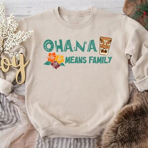 May include: Beige sweatshirt with the text "OHANA MEANS FAMILY" in teal and orange. The design includes a tiki mask and colorful flowers. A cozy, casual top for family gatherings or everyday wear.