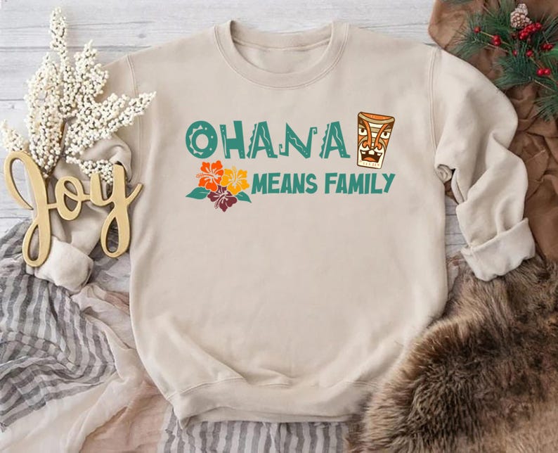 May include: Beige sweatshirt with the text "OHANA MEANS FAMILY" in teal and orange. The design includes a tiki mask and colorful flowers. A cozy, casual top for family gatherings or everyday wear.