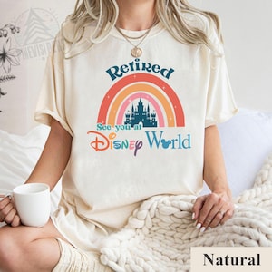 K&ouml;nnte beinhalten: Ein wei&szlig;es T-Shirt mit einem Regenbogen- und Schloss-Motiv. Der Text "Retired See you at Disney World" ist auf dem Shirt aufgedruckt.