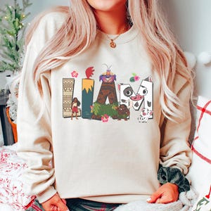 May include: Beige sweatshirt featuring the word "IAM" in colourful, cartoon lettering. The letters are decorated with images of characters and elements from a popular animated film. The sweatshirt has a crew neck and long sleeves.