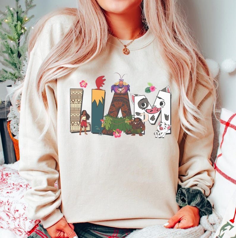 May include: Beige sweatshirt featuring the word "IAM" in colourful, cartoon lettering. The letters are decorated with images of characters and elements from a popular animated film. The sweatshirt has a crew neck and long sleeves.