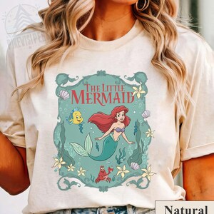 May include: A cream-colored t-shirt featuring a teal framed graphic of Ariel from The Little Mermaid. The graphic includes Ariel with red hair and a green tail, a yellow fish, and a red crab. The words "The Little Mermaid" are at the top.