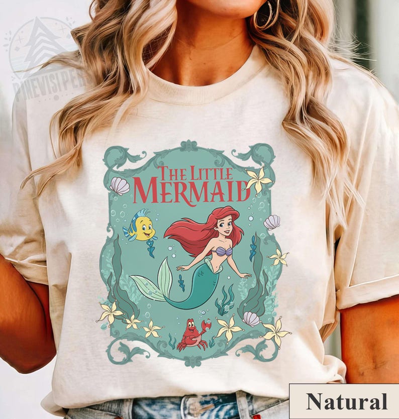 May include: A cream-colored t-shirt featuring a teal framed graphic of Ariel from The Little Mermaid. The graphic includes Ariel with red hair and a green tail, a yellow fish, and a red crab. The words "The Little Mermaid" are at the top.