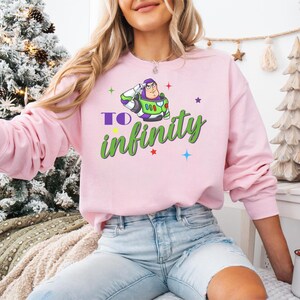 May include: A light pink sweatshirt featuring a graphic of Buzz Lightyear saluting, with the words "TO infinity" in green and purple. The sweatshirt has long sleeves and a crew neck.
