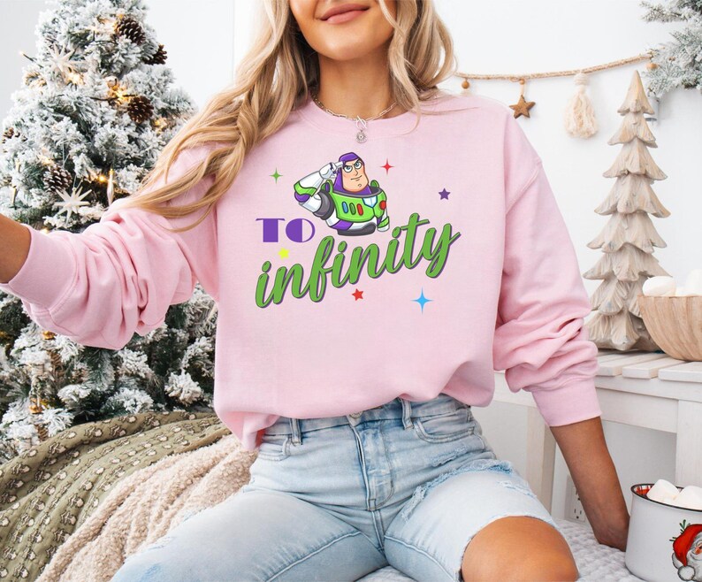 May include: A light pink sweatshirt featuring a graphic of Buzz Lightyear saluting, with the words "TO infinity" in green and purple. The sweatshirt has long sleeves and a crew neck.