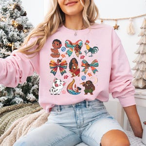 May include: A light pink sweatshirt featuring colorful cartoon illustrations of characters and bows. The design includes a baby, a rooster, a pig, and other figures. The sweatshirt is long-sleeved and has a crew neck.