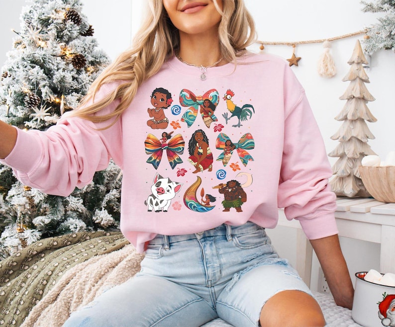 May include: A light pink sweatshirt featuring colorful cartoon illustrations of characters and bows. The design includes a baby, a rooster, a pig, and other figures. The sweatshirt is long-sleeved and has a crew neck.