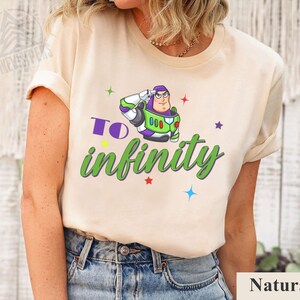 May include: A beige t-shirt featuring a graphic of Buzz Lightyear saluting, with the words "To infinity" in green and purple. Colorful stars surround the graphic. The shirt is made of a soft material.