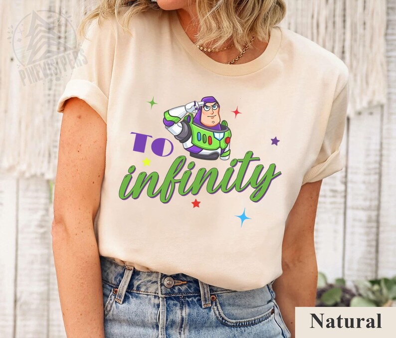May include: A beige t-shirt featuring a graphic of Buzz Lightyear saluting, with the words "To infinity" in green and purple. Colorful stars surround the graphic. The shirt is made of a soft material.
