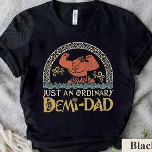 Op de afbeelding: Zwart T-shirt met een cartoon afbeelding van een gespierde man met een kroon en de tekst "Just an ordinary Demi-Dad" in een vintage lettertype.