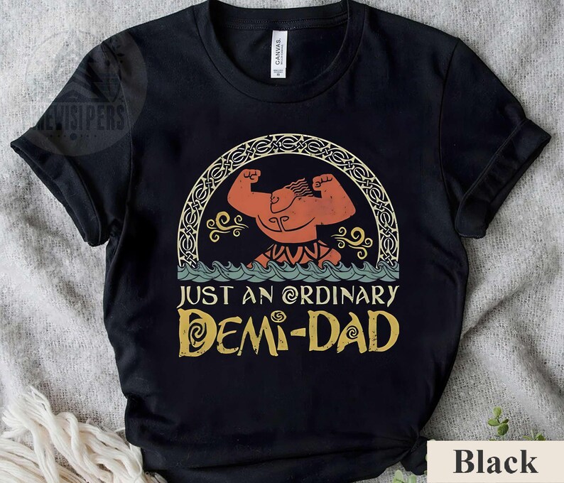 Op de afbeelding: Zwart T-shirt met een cartoon afbeelding van een gespierde man met een kroon en de tekst "Just an ordinary Demi-Dad" in een vintage lettertype.