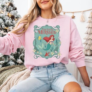 May include: A light pink sweatshirt featuring a graphic of Ariel from The Little Mermaid. The design includes Ariel, Flounder, Sebastian, and the words "The Little Mermaid" in a decorative frame. The sweatshirt has long sleeves.