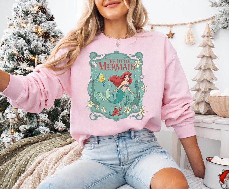 May include: A light pink sweatshirt featuring a graphic of Ariel from The Little Mermaid. The design includes Ariel, Flounder, Sebastian, and the words "The Little Mermaid" in a decorative frame. The sweatshirt has long sleeves.