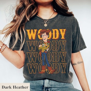 Puede incluir: Camiseta gris oscuro con un dise&ntilde;o del personaje Woody de Toy Story. El dise&ntilde;o incluye la palabra "WOODY" en naranja y el personaje con un atuendo de vaquero. La camiseta es de manga corta y cuello redondo.