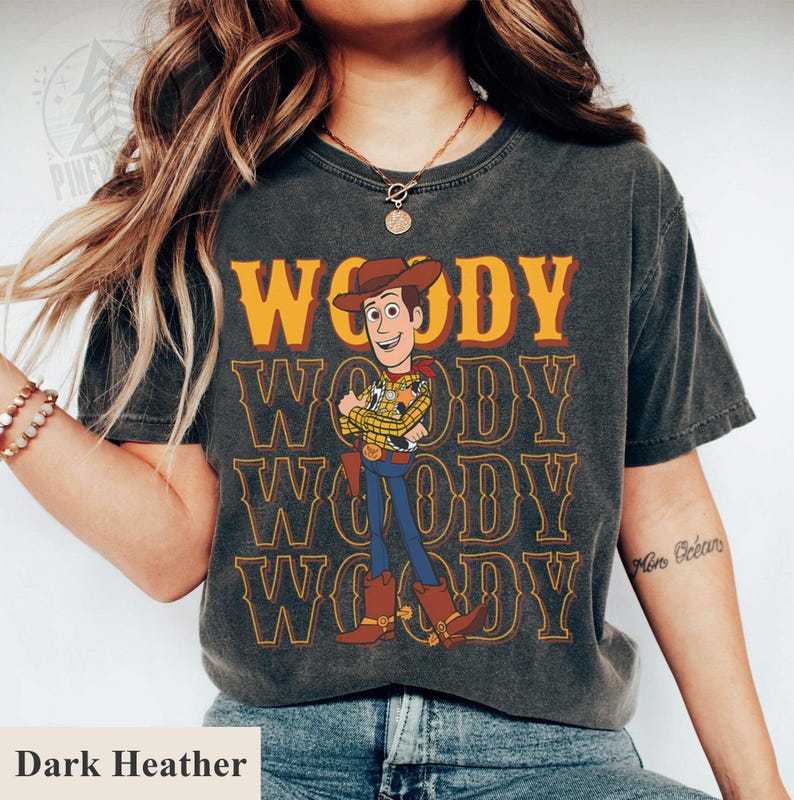 Puede incluir: Camiseta gris oscuro con un dise&ntilde;o del personaje Woody de Toy Story. El dise&ntilde;o incluye la palabra "WOODY" en naranja y el personaje con un atuendo de vaquero. La camiseta es de manga corta y cuello redondo.