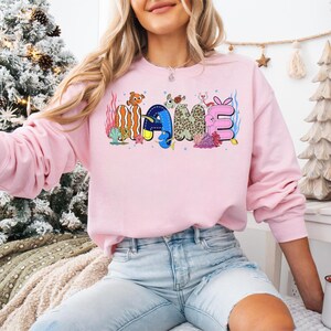 May include: A light pink sweatshirt with the word "NAME" in colorful, cartoon-style lettering. The letters are decorated with marine life and coral. The sweatshirt has long sleeves and a crew neck.