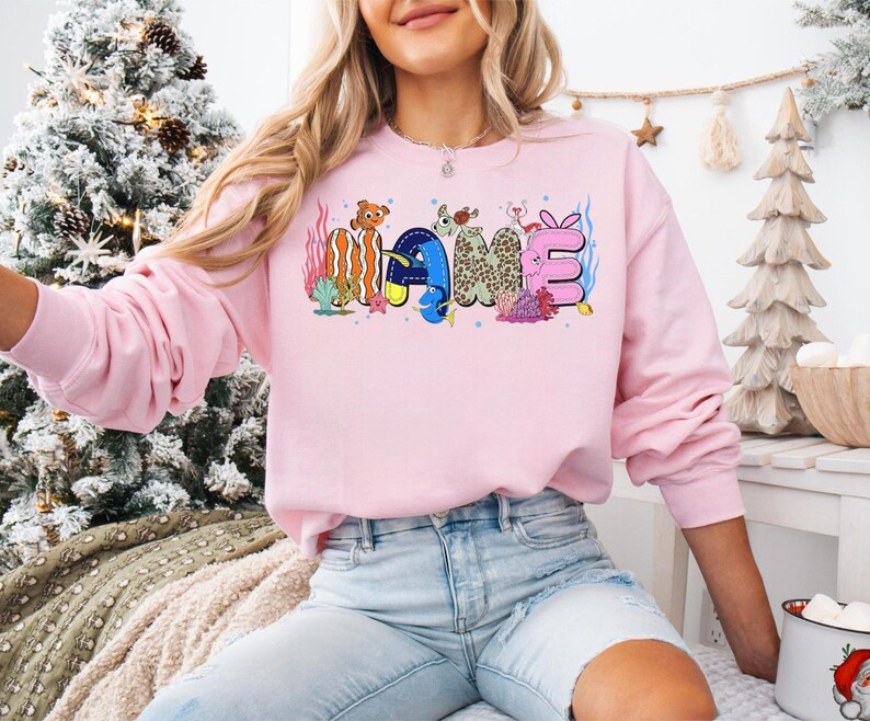 May include: A light pink sweatshirt with the word "NAME" in colorful, cartoon-style lettering. The letters are decorated with marine life and coral. The sweatshirt has long sleeves and a crew neck.